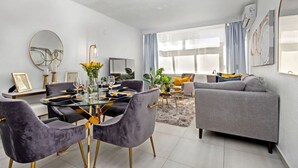 A modern living room with a glass dining table, grey upholstered chairs, a grey sofa, and a large window with curtains.