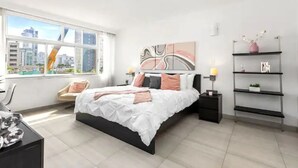 A modern bedroom with a large bed, a desk, and a chair. There is a window with a city view.