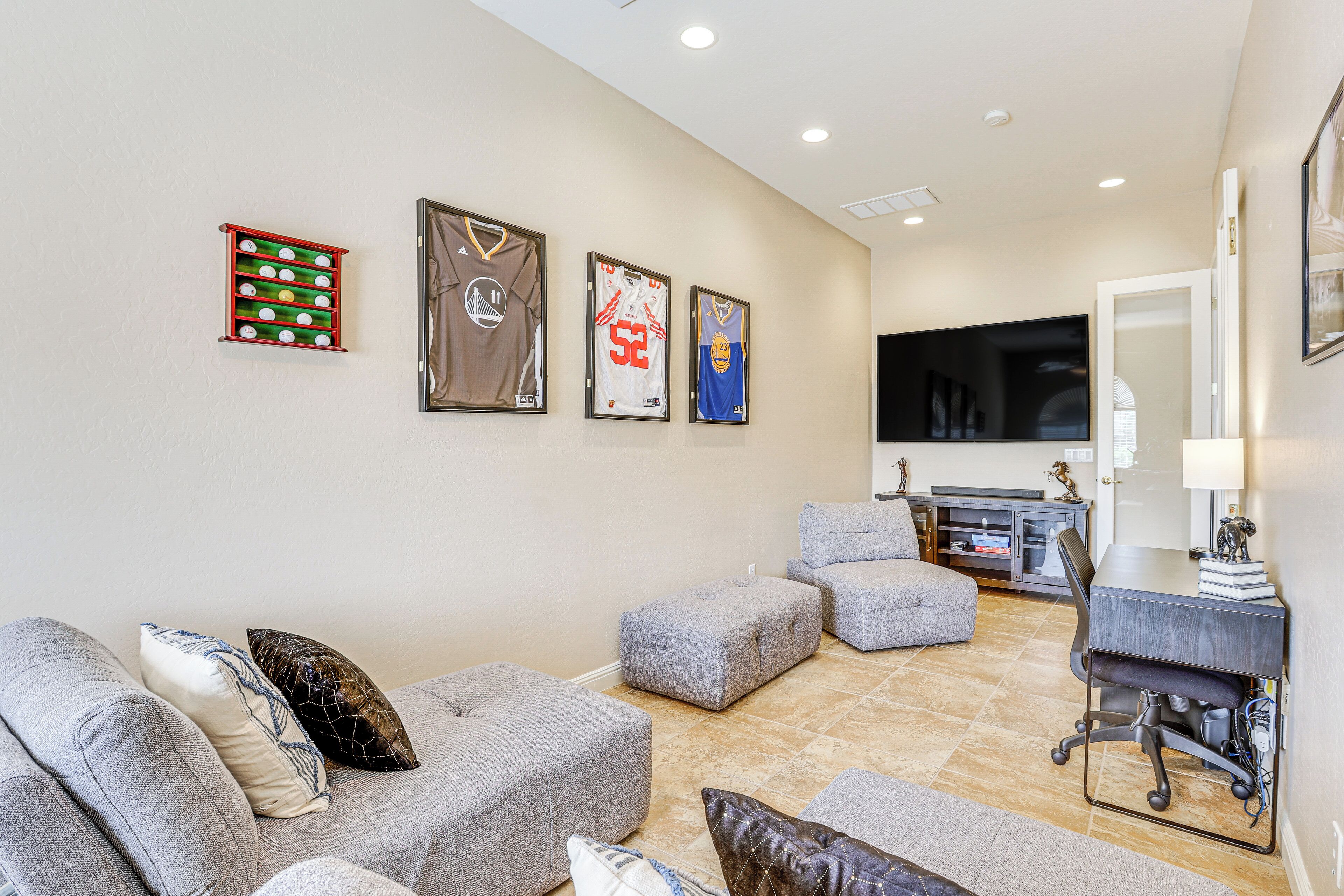 A living room with grey sofas, ottomans, and a flat-screen TV. There are framed sports jerseys on the wall.