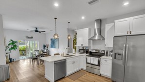 A modern kitchen with white cabinets, stainless steel appliances, and a dining area with a view to the outdoors. - NEW Luxury pool home w/hot tub, KING bed, close to beach, firepit (St. Petersburg)