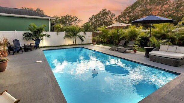 A rectangular swimming pool with lounge chairs and umbrellas, surrounded by greenery and a white fence. - NEW Luxury pool home w/hot tub, KING bed, close to beach, firepit (St. Petersburg)
