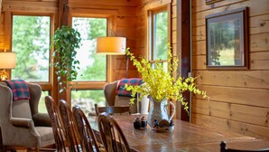 A wooden cabin interior with a dining table, chairs, a lamp, a vase with yellow flowers, and framed pictures on the walls. - Poconos True Log Cabin with Views, Hot Tub & Game Room (Mount Pocono)