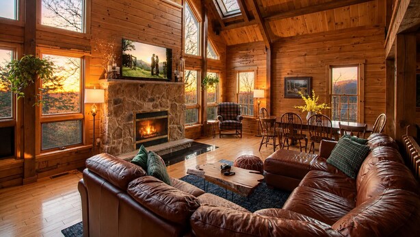 A log cabin interior with a stone fireplace, leather furniture, and large windows. - Poconos True Log Cabin with Views, Hot Tub & Game Room (Mount Pocono)