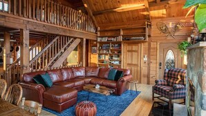 A cozy cabin interior with a wooden staircase, a leather sectional sofa, a plaid armchair, and a bookshelf. - Poconos True Log Cabin with Views, Hot Tub & Game Room (Mount Pocono)