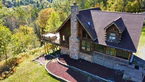 A log cabin with a stone chimney, large windows, and a deck overlooking a forested area. - Poconos True Log Cabin with Views, Hot Tub & Game Room (Mount Pocono)