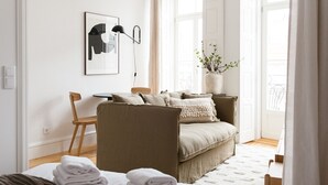 Ett modernt och väl upplyst rum med en soffa, en fåtölj, ett bord och en säng med prydligt hopvikta handdukar.