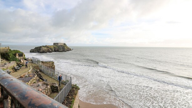 A coastal view with a fort on a rocky outcrop, a walkway along the shore, and a clear sky. - 1 St. Marys Court (Tenby)