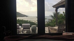 A bedroom with a large window overlooking a beach and ocean. There are two chairs and a table outside the window.