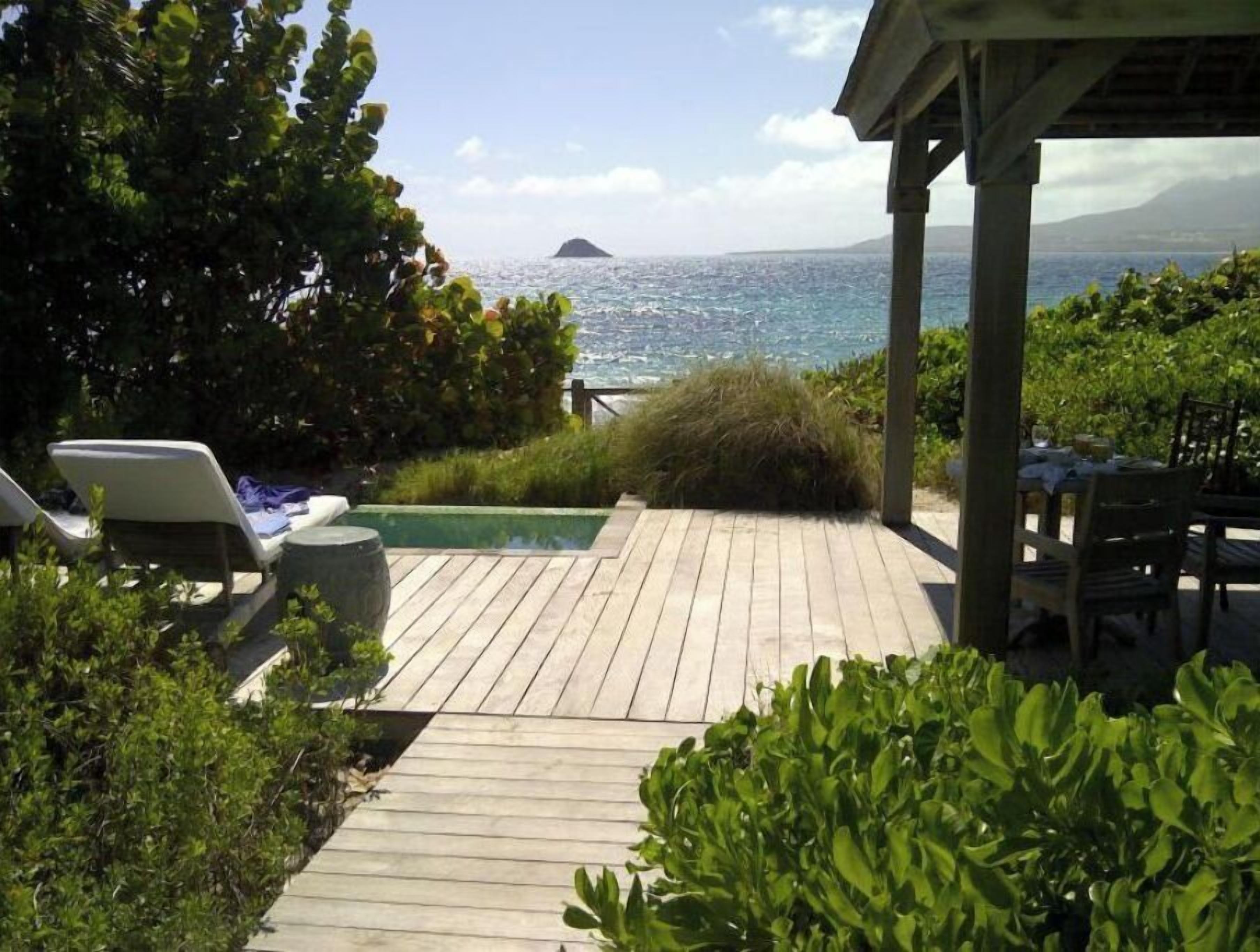 Eine Holzterrasse mit Liegestühlen und einem kleinen Pool mit Blick auf ein Gewässer und eine entfernte Insel.