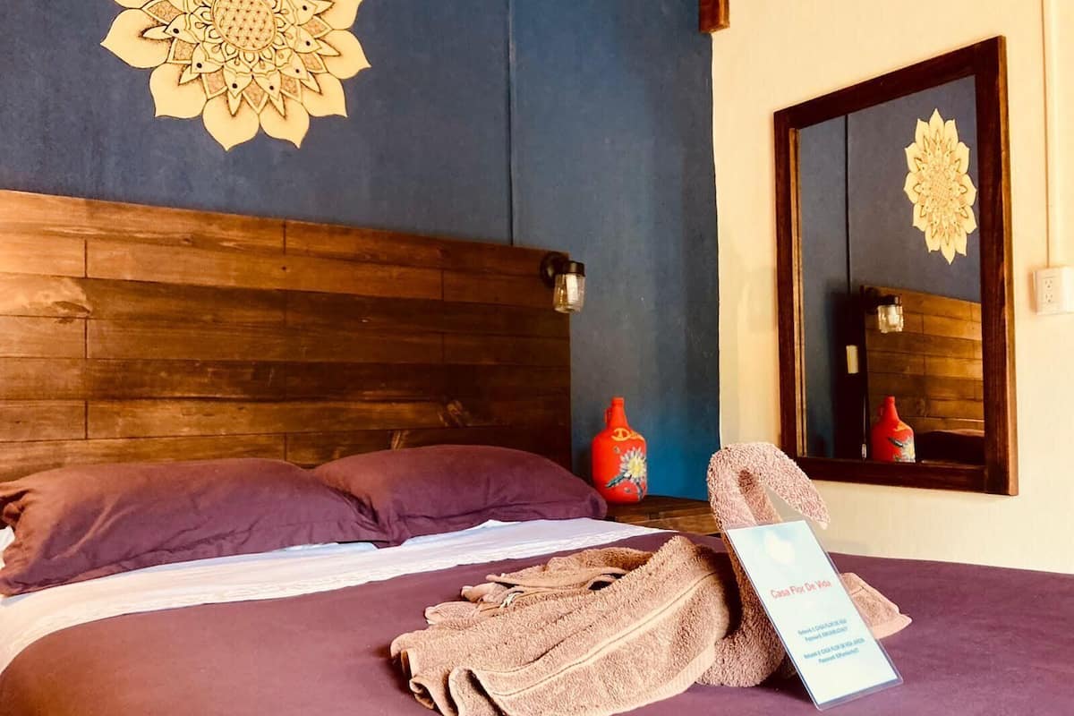 A neatly made bed with a purple bedspread and two pillows, a wooden headboard, a large mirror with a decorative frame, and a sign on the bed.
