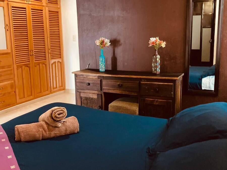 A bedroom with a bed, a wooden wardrobe, a desk with two vases, and a mirror.