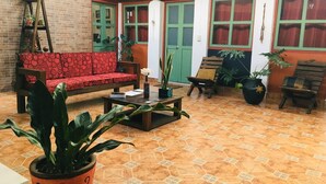 Ein gemütlicher Innenhof mit einem rot gemusterten Sofa, einer hölzernen Bank und einem Couchtisch. Mehrere Blumentöpfe und eine an der Wand lehnende Leiter sind ebenfalls vorhanden.