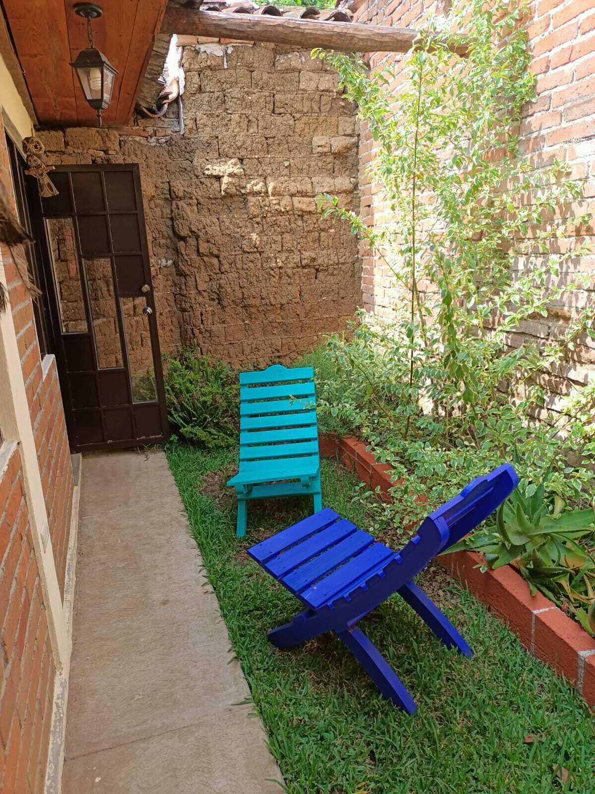 Un estrecho sendero conduce a una pequeña zona de estar al aire libre con un banco y una silla reclinable, rodeada de un muro de ladrillo y vegetación.