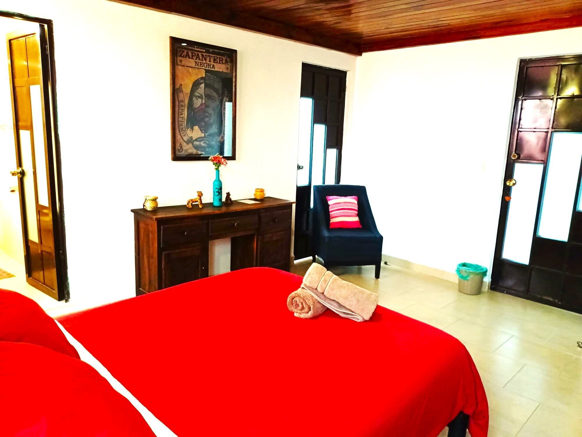 Une chambre d’hôtel avec un lit rouge, une commode en bois, une chaise et une affiche au mur.