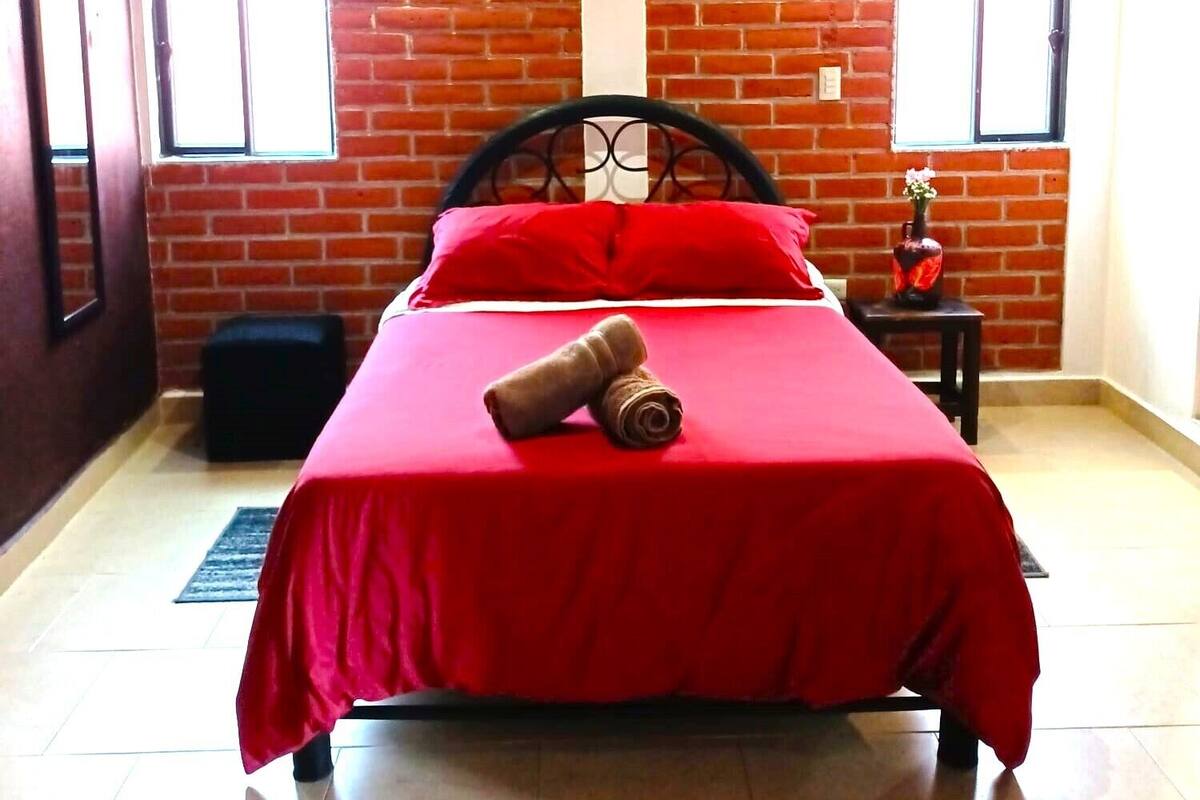 A room with a brick wall, a bed with a red bedspread, a wooden ceiling, and two windows.