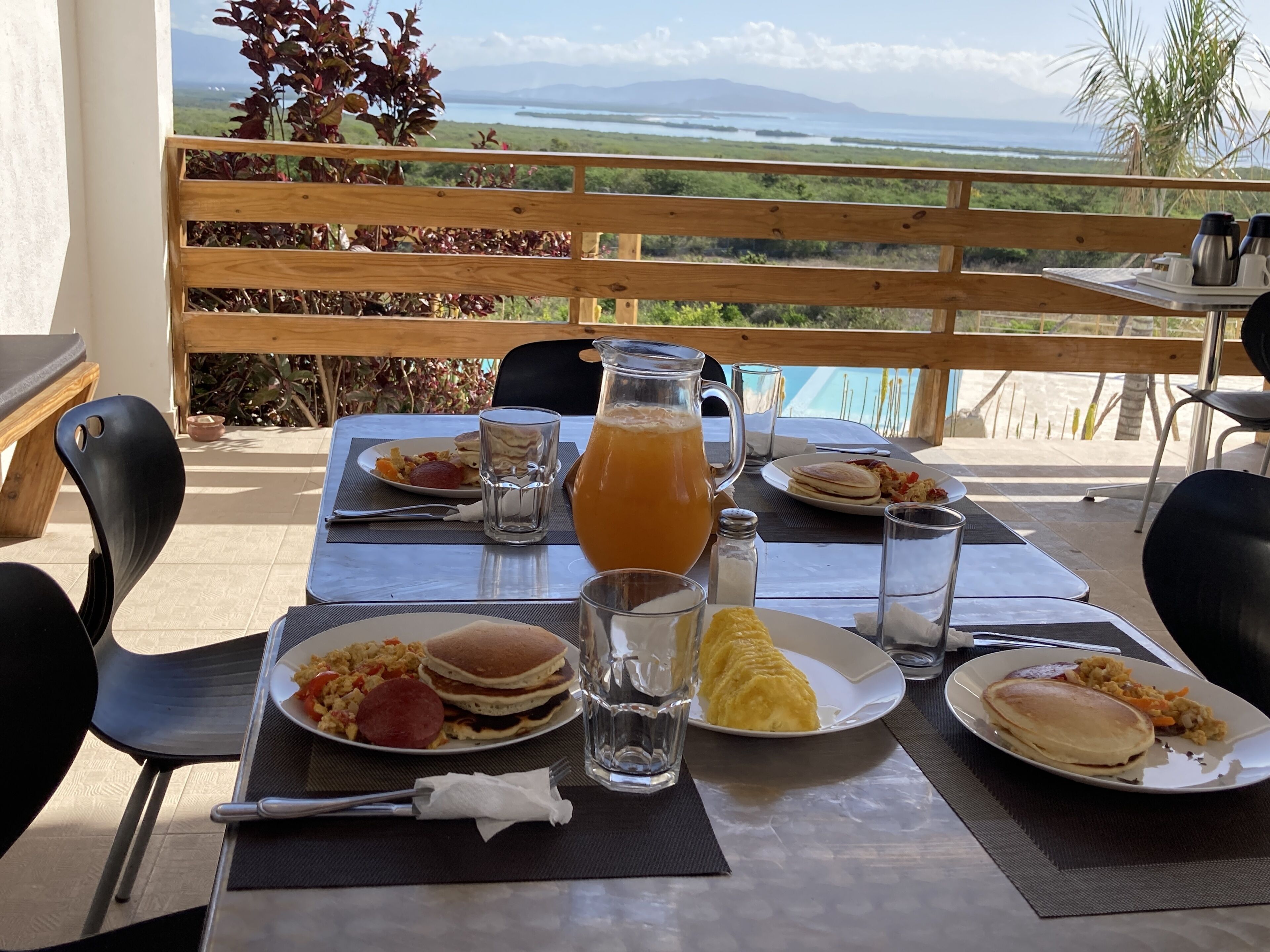 Una tavola imbandita per la colazione, con diversi piatti, una brocca di succo d'arancia e vista sull'oceano e sulle montagne.