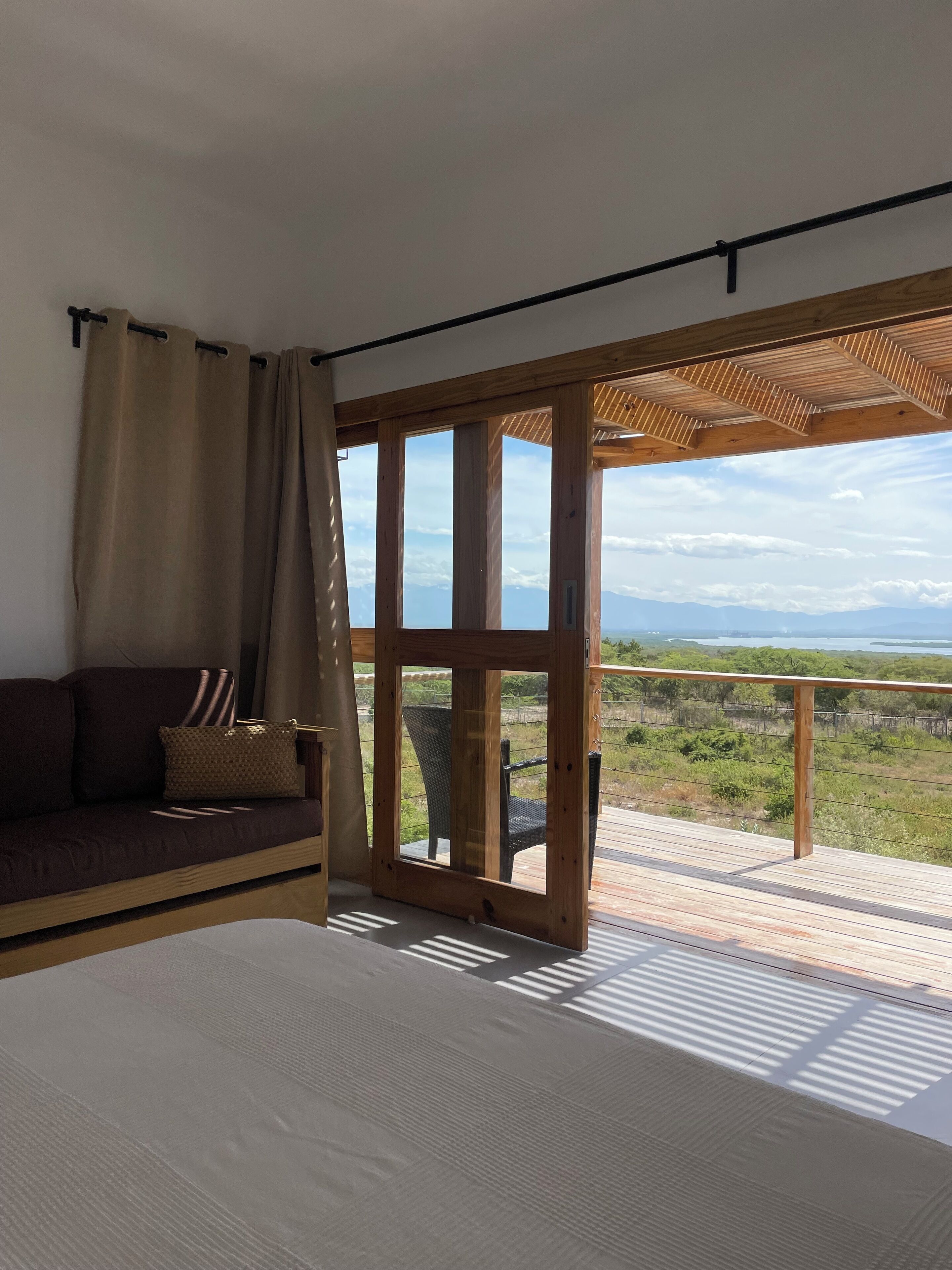 A bedroom with a large bed, a wooden ceiling, a view of the outdoors, and a balcony with a chair.