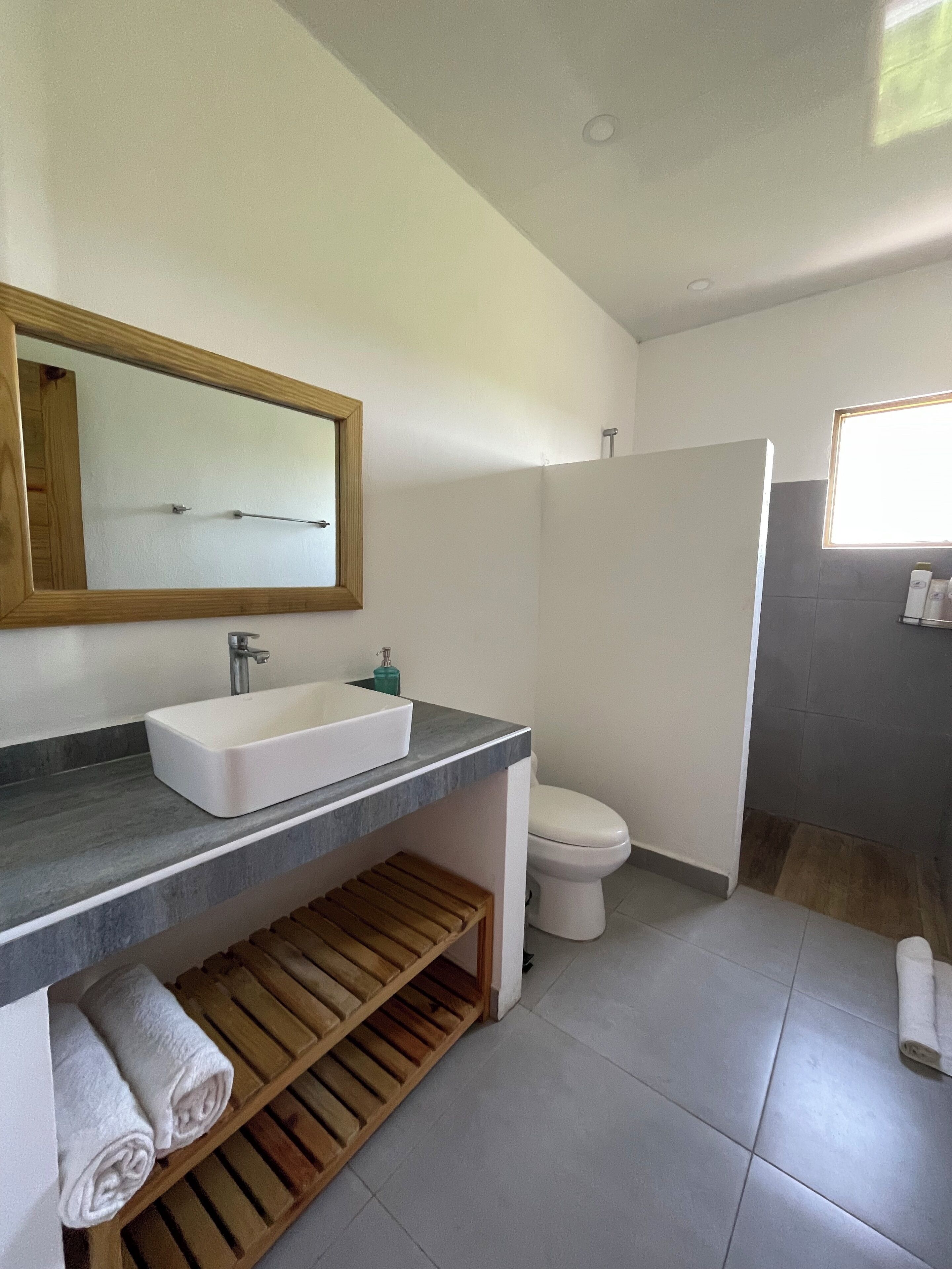 Un baño moderno con un lavamanos blanco, un estante de madera, un inodoro y una zona de ducha.