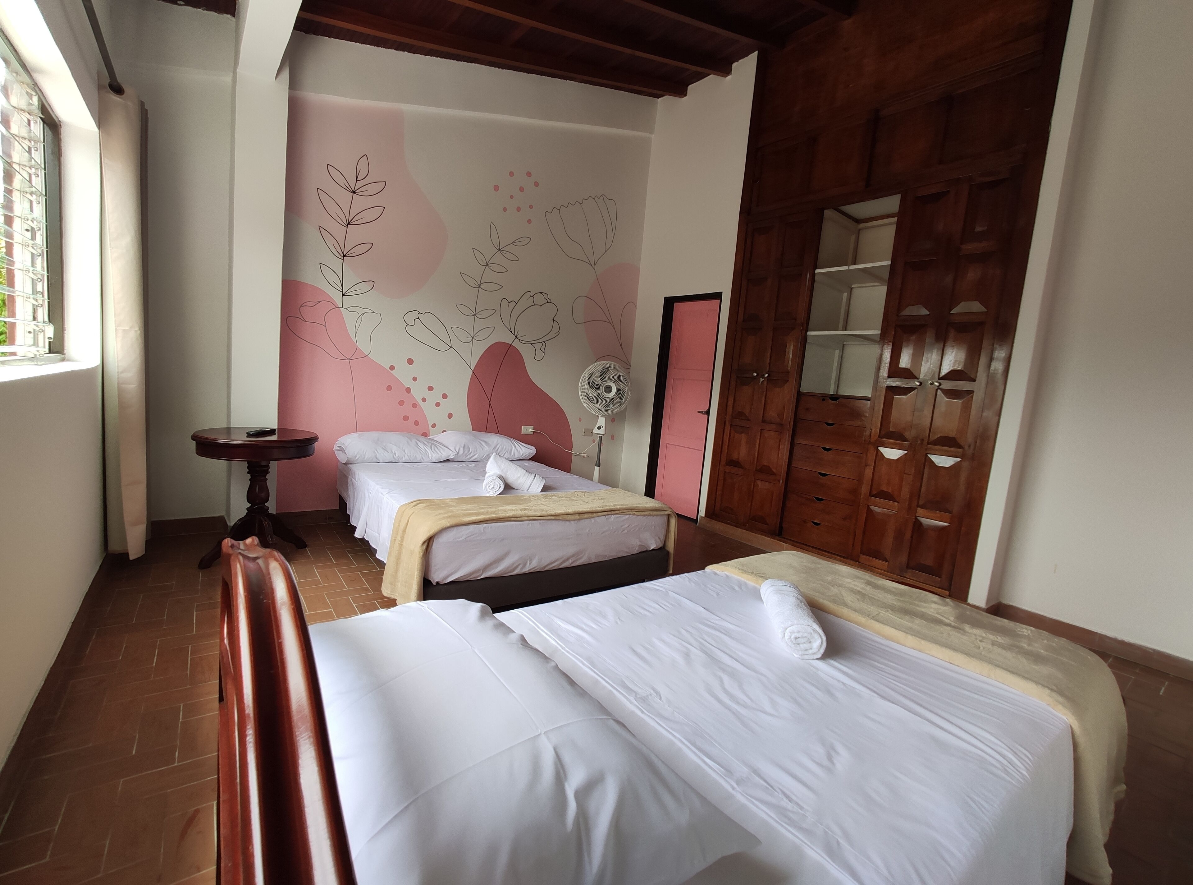 Quarto de hotel com duas camas, um guarda-roupa de madeira, uma mesa pequena e um painel decorativo.