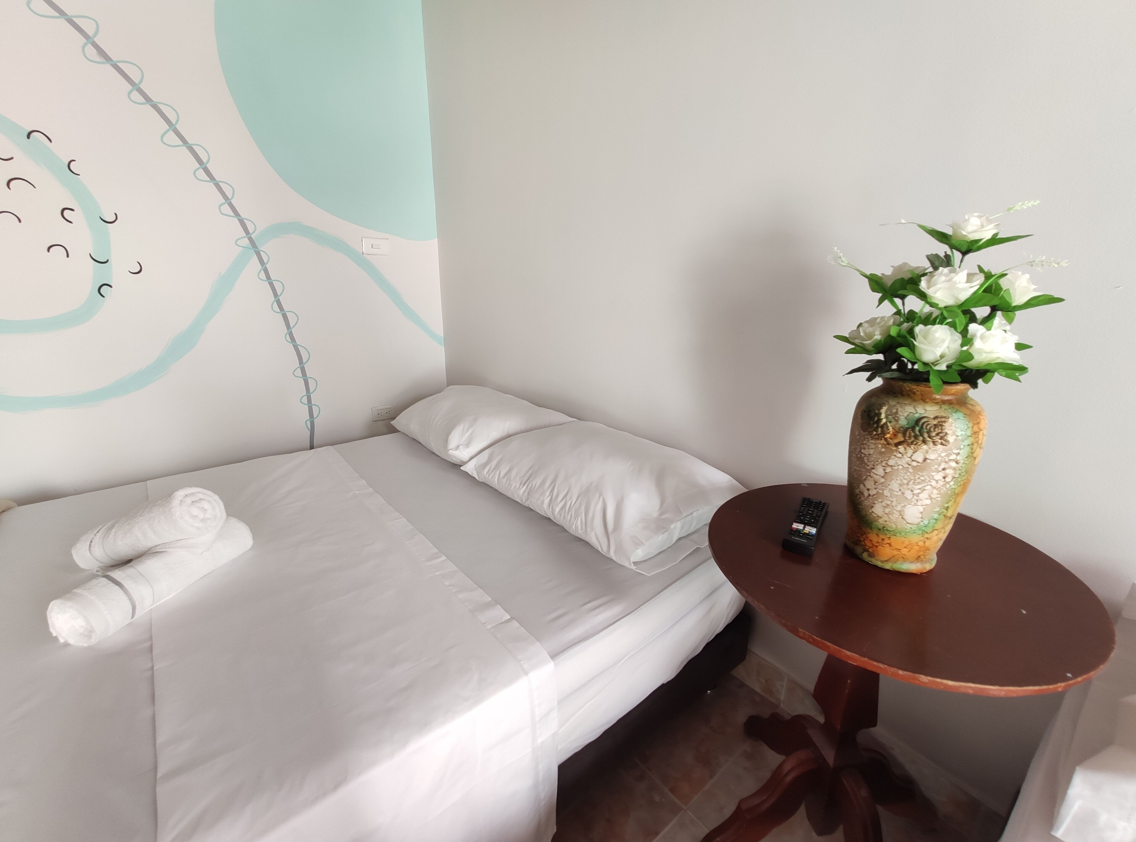 Una cama bien hecha con sábanas blancas, un florero con flores blancas sobre una mesita redonda y una pared con un diseño sencillo.