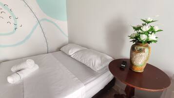 Una cama bien hecha con sábanas blancas, un florero con flores blancas sobre una mesita redonda y una pared con un diseño sencillo.