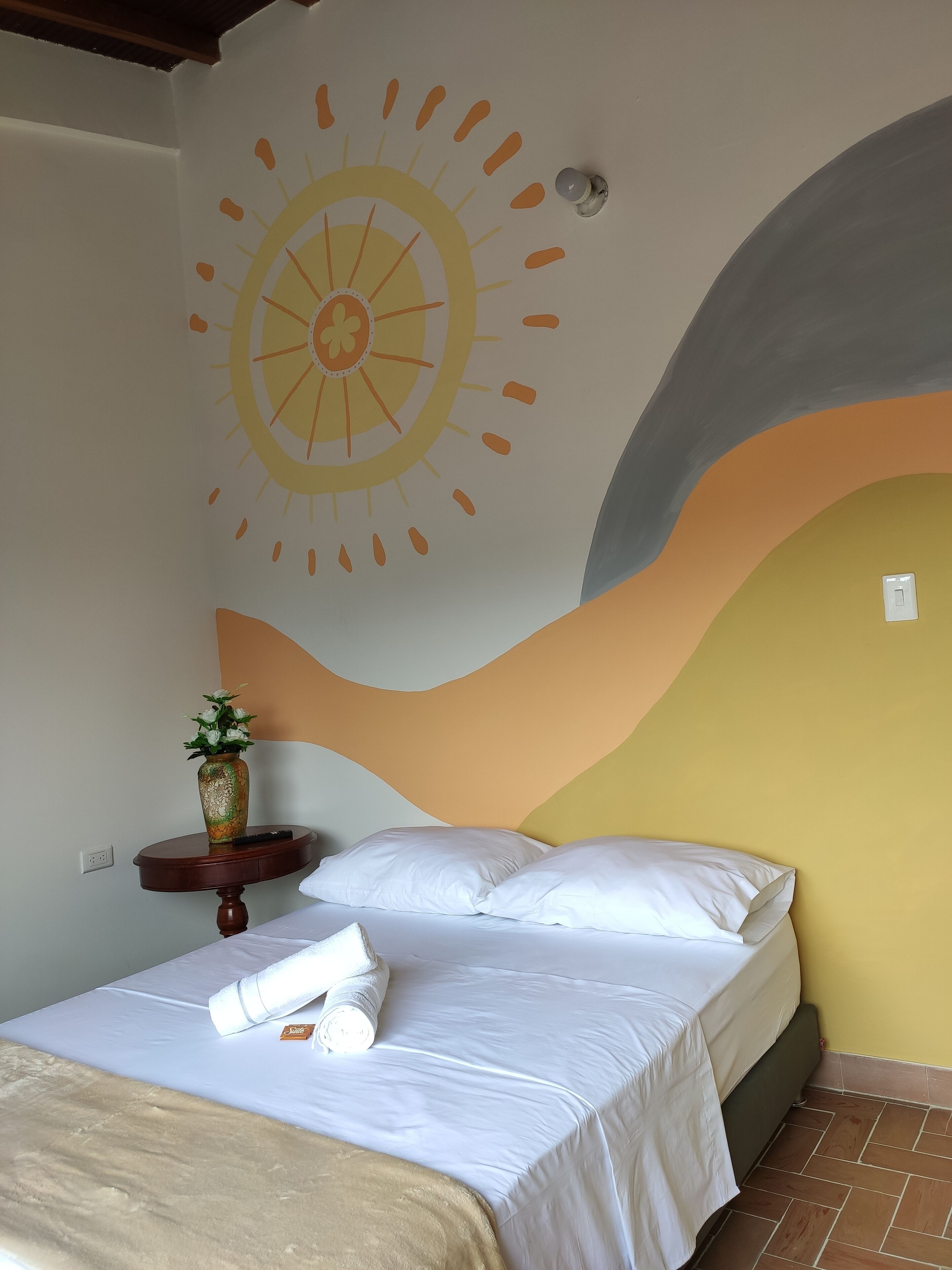 A bedroom with a bed, pillows, a vase with flowers, and a wall with a sun design.