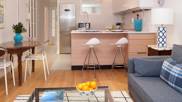 Una cocina moderna con una isla central, taburetes de bar, una mesa de comedor con sillas, un sofá y una mesa de centro con un cuenco de naranjas.