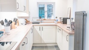A modern kitchen with white cabinets, a sink, a window with a view of trees, and various kitchen appliances. - Beautiful, 17th century Lake District cottage close to village and mountains (Coniston)