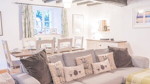 A cozy living room with a grey sofa, a dining table set for four, and a window with curtains. - Beautiful, 17th century Lake District cottage close to village and mountains (Coniston)