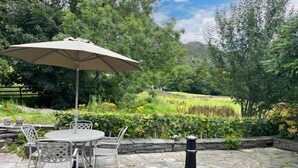 An outdoor patio with a stone table and chairs, a metal grill, and an umbrella. - Beautiful, 17th century Lake District cottage close to village and mountains (Coniston)