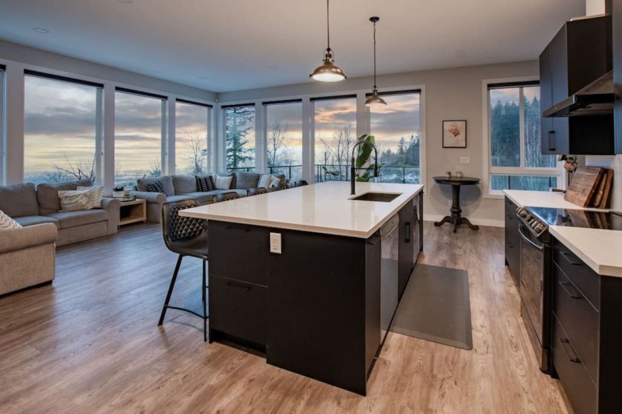 A modern kitchen with a large island, wooden flooring, and large windows offering views of the outdoors.