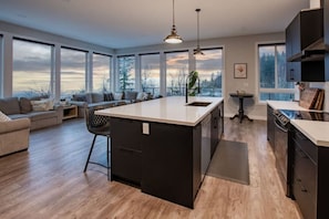 A modern kitchen with a large island, wooden flooring, and large windows offering views of the outdoors.