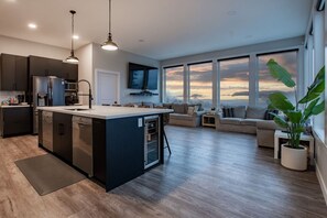 A modern kitchen with a central island, a dining area with a sofa, and large windows offering a view of the sky.