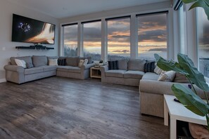 A modern living room with a large flat-screen TV, two grey sofas, a coffee table, and large windows offering a view of the sky. - #TheMonopolyHouse-Private acreage /Bar/Arcade/Gym/HotTub/Sauna/Sports court/golf (Chilliwack)