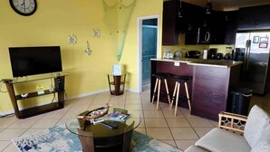 A living room with a TV, coffee table, and sofa, a kitchen with a fridge and bar stools, and a yellow wall with decorative items. - Relaxing 1Bed 1Bath Villa In Downtown Red Hook! (Saint Thomas)