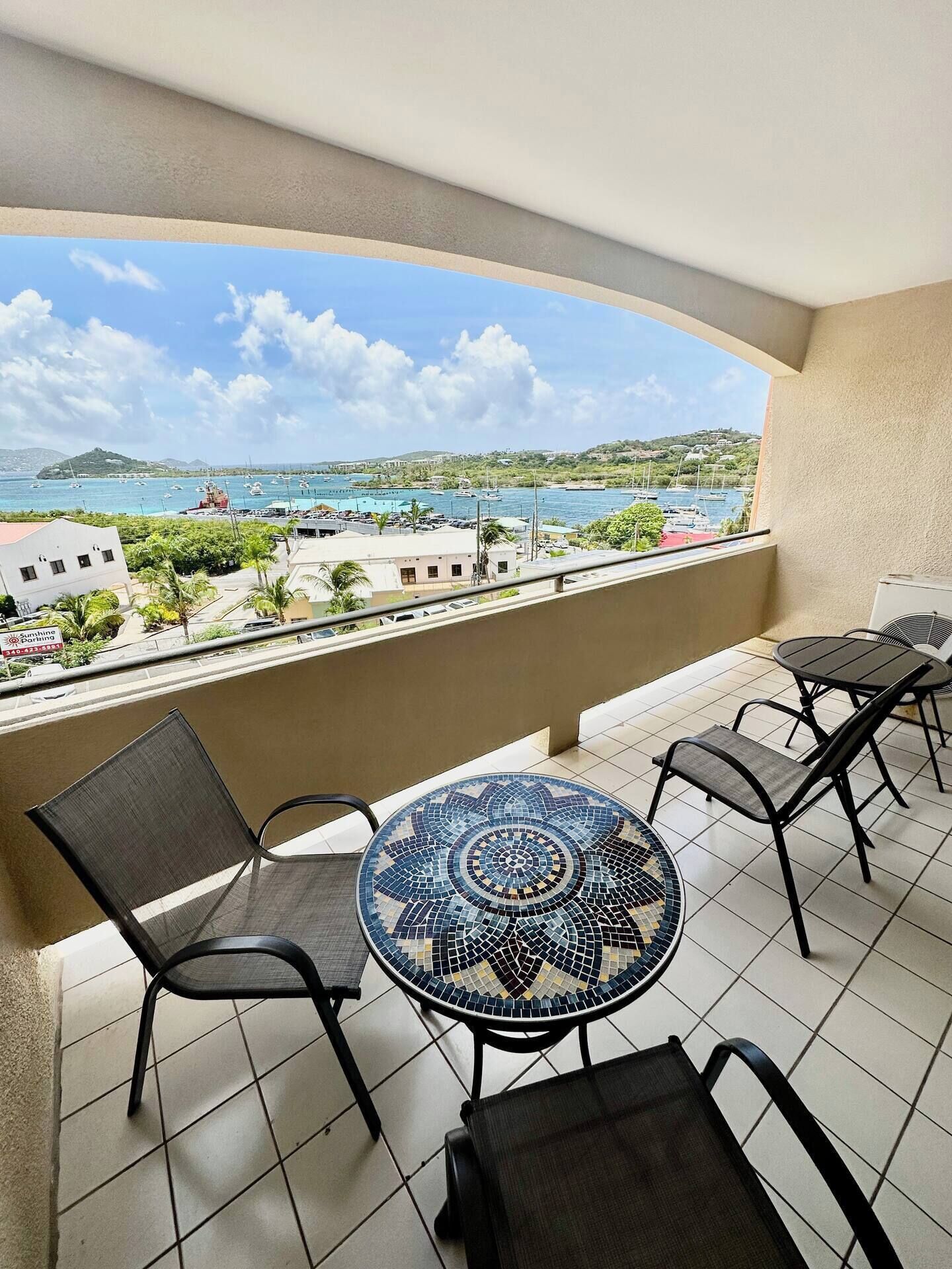 A balcony with a tiled floor, a round table with chairs, and a view of a coastal area with boats and buildings.