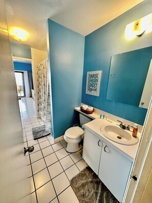 Une salle de bain avec des toilettes, un lavabo et un miroir. Il y a une affiche sur le mur qui dit : « Soleil levant, coups de soleil, coucher de soleil, et ça recommence. »