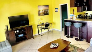 Una sala de estar con televisión, sofá, mesa de centro y una cocina al fondo.