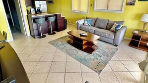 Sala de estar con sofá, mesa de centro y ventilador de techo. Hay una cocina con refrigerador y mesa de comedor.
