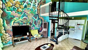 A living room with a colorful mural, a sofa, a coffee table, a TV, and a staircase. - Harbor View: 2Bed 1Bath Loft In Downtown Red Hook! (East End)