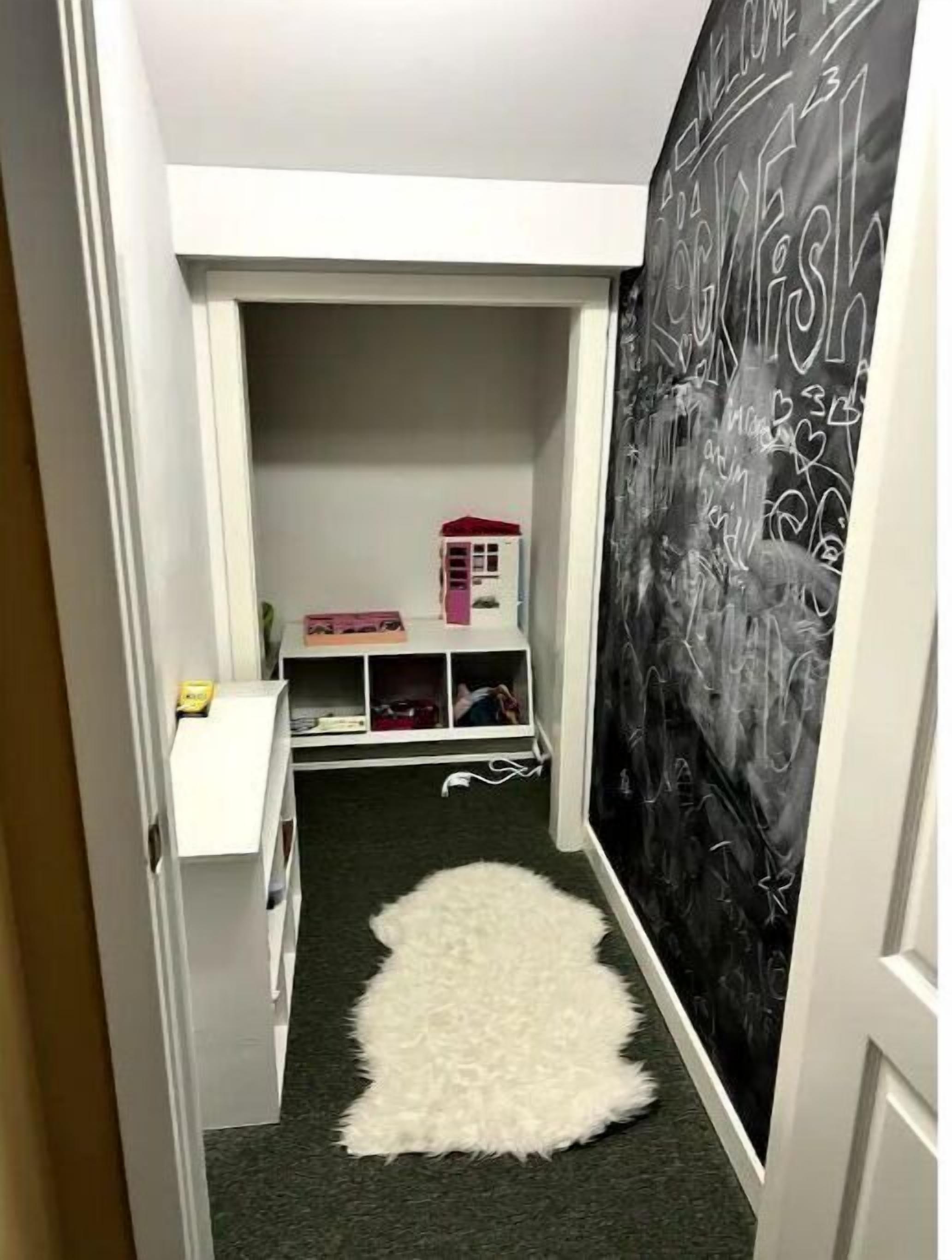 Un pasillo estrecho con un manto blanco de piel, una pequeña casa de juguete rosa y una pizarra con la palabra 'WELCOME' escrita.