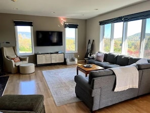 A spacious living room with a large grey sofa, a television, and a view of the outdoors. - Amazing Mountain Views, Hot Tub and Close to Wineries and Breweries (Afton)