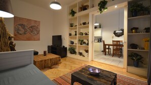 Ein Wohnzimmer mit einer Couch, einem Holztisch und einem Fernseher. Regale mit Pflanzen und ein Essbereich mit Stühlen sind ebenfalls vorhanden.
