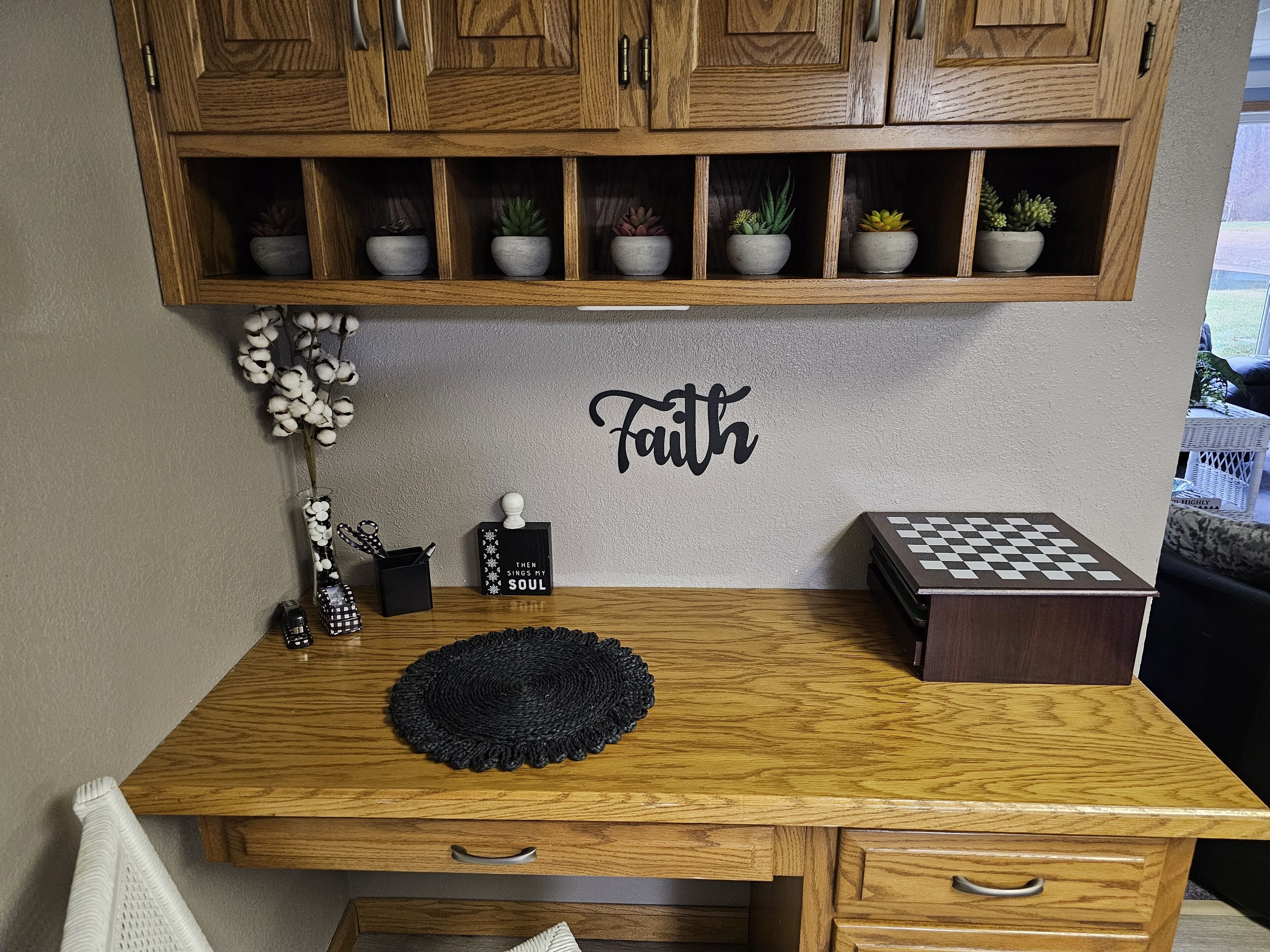 Un bureau en bois surmonté d’un set de table en fibres tressées, un jeu d’échecs, un élément décoratif mural portant le mot «Foi», et une étagère avec des plantes en pot.