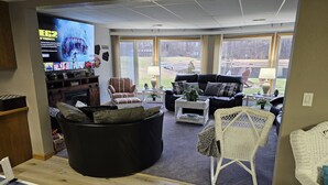 Una sala de estar con un sofá seccional negro, un sillón a rayas y una silla de mimbre blanca. Hay un televisor de pantalla plana montado en la pared, y un ventanal ofrece vistas al exterior.