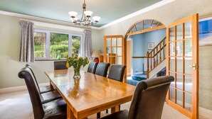 A dining room with a wooden table and chairs, a chandelier, and a view of a garden through the windows. - Seahaven - Luxury Cottages (Torquay)