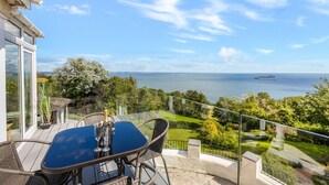 A balcony with a glass table and chairs, overlooking a scenic view of the sea and a yacht. - Seahaven - Luxury Cottages (Torquay)