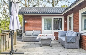 Een houten terras met twee rieten banken en een tafel, een overdekte parasol, en een huis met houten bekleding en een pannendak.