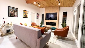 A modern living room with a grey sofa, orange and brown chairs, a wooden ceiling, and a TV mounted on the wall.