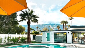 A modern outdoor pool area with a view of mountains, surrounded by palm trees and a white fence.